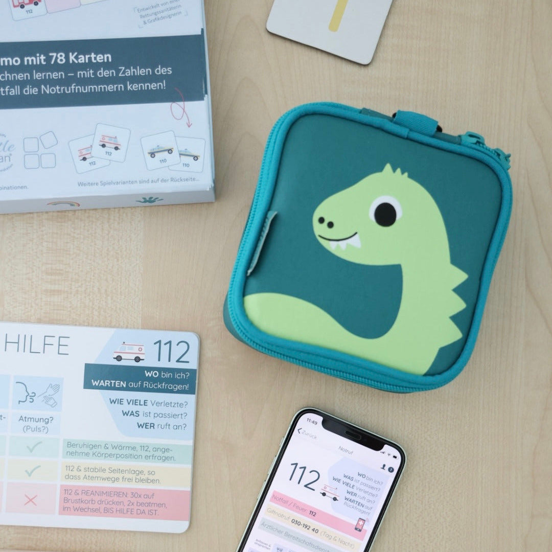 littlebag Erste-Hilfe-Tasche im Dino-Design neben Erste-Hilfe-Plan, Notruf-Memo-Spiel und Smartphone mit der littleplan Erste-Hilfe-App
