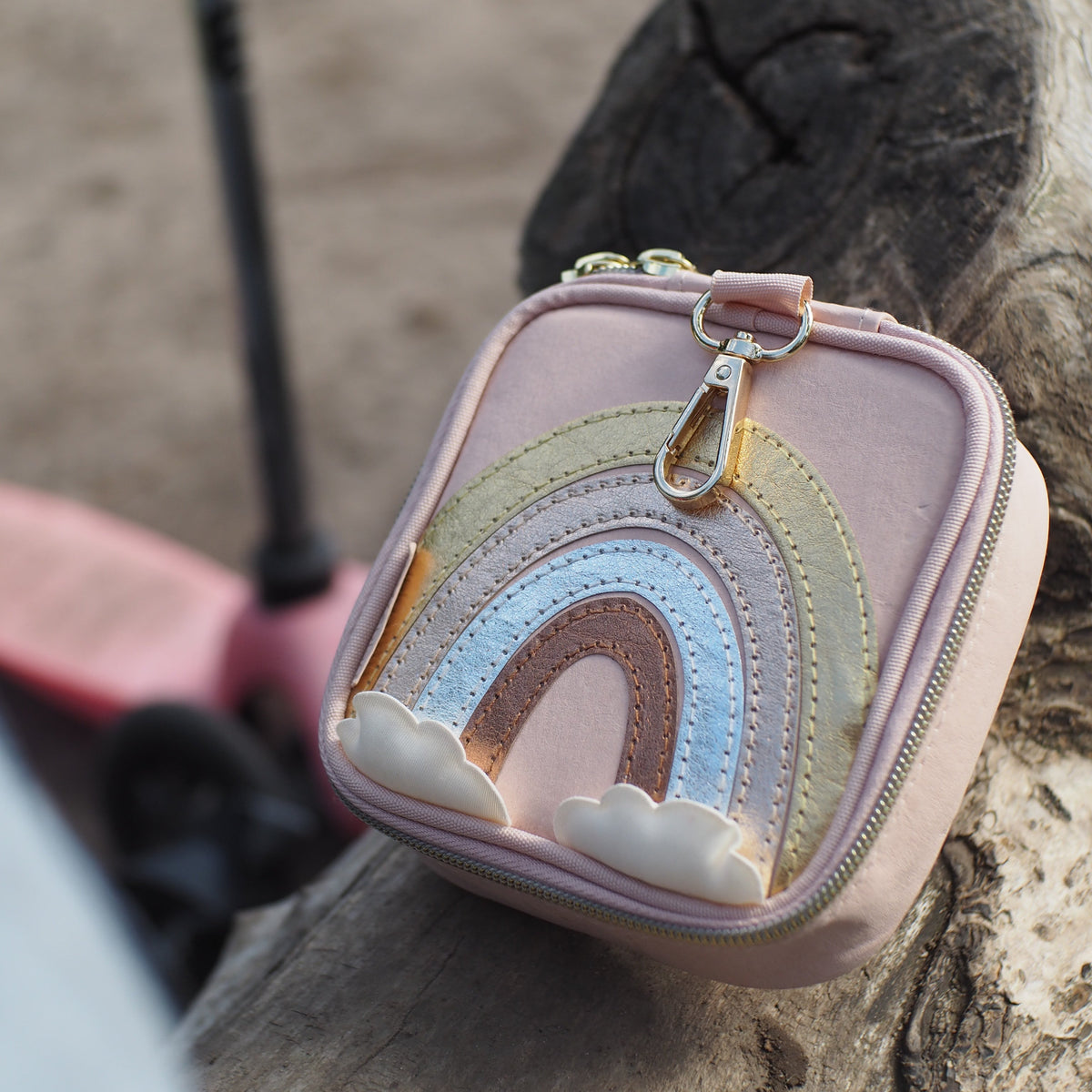 Nahaufnahme der littlebag Erste-Hilfe-Tasche im Regenbogen-Design mit goldenem Karabinerhaken auf einem Baumstamm, im Hintergrund ein Kinderroller